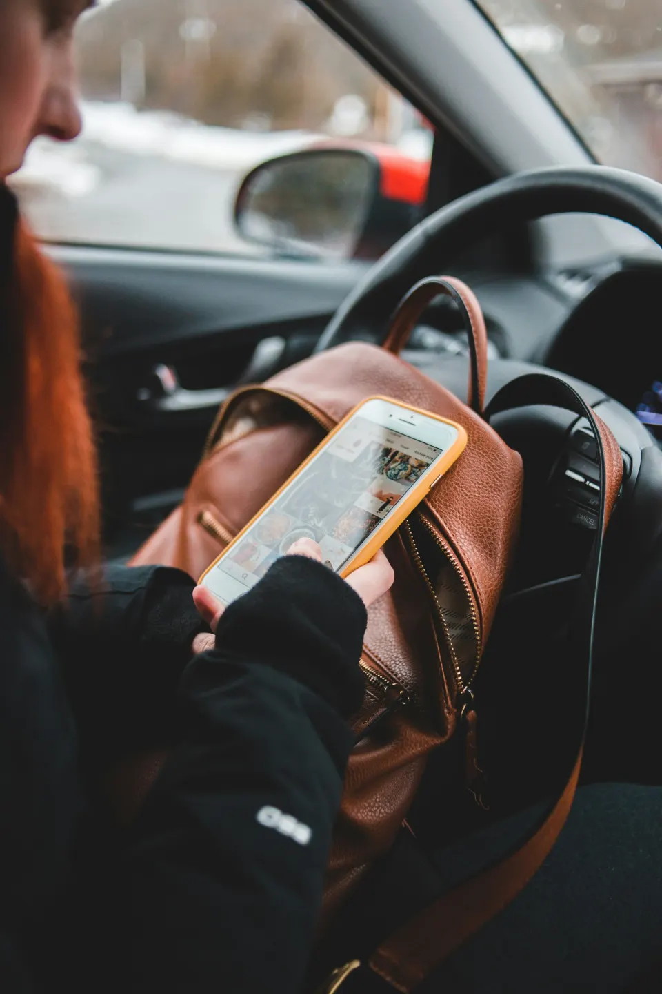 Person with long red hair sitting in car, looking at phone, with a brown purse on lap and dashboard in background.