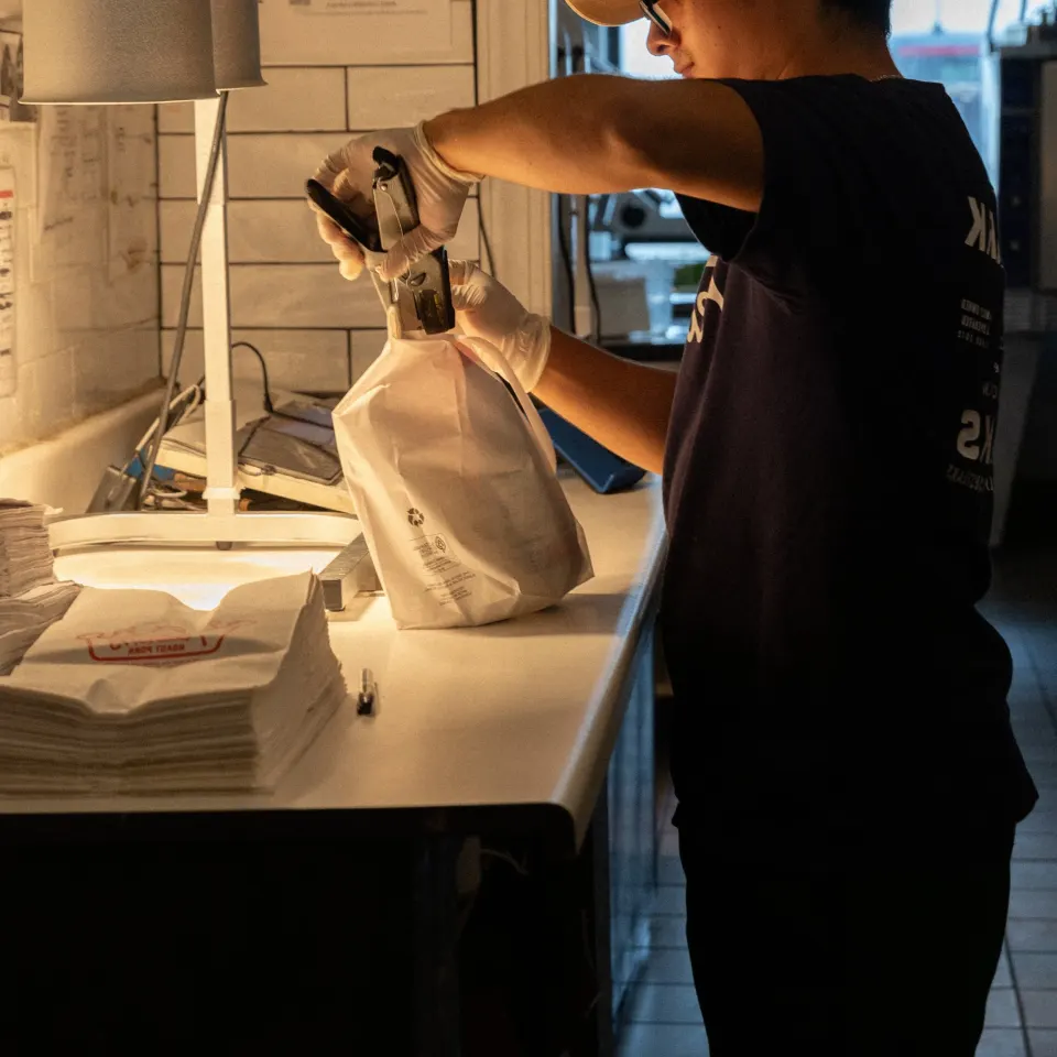 A person wearing gloves is sealing a paper bag at a counter in a restaurant or cafe, with stacks of paper bags nearby.